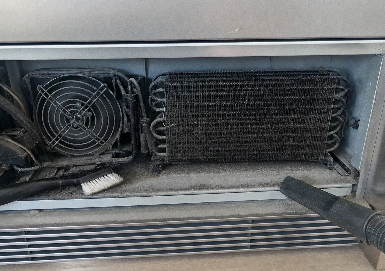 Condenser coil and fan behind a removed lower grille, with a vacuum nozzle and brush against the fins